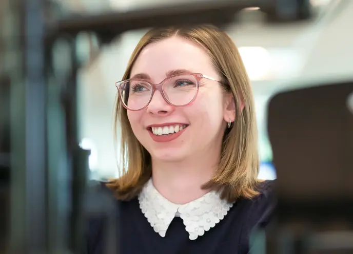 A woman wearing glasses smiling at camera whilst typing at a computer screen in an office setting.